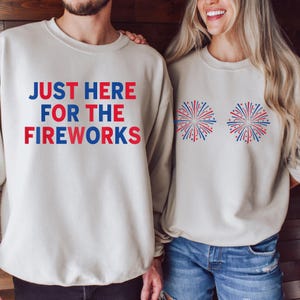 May include: Two beige crewneck sweatshirts. The left sweatshirt features the text "JUST HERE FOR THE FIREWORKS" in alternating red and blue block letters. The right sweatshirt features two graphic illustrations of red and blue fireworks.