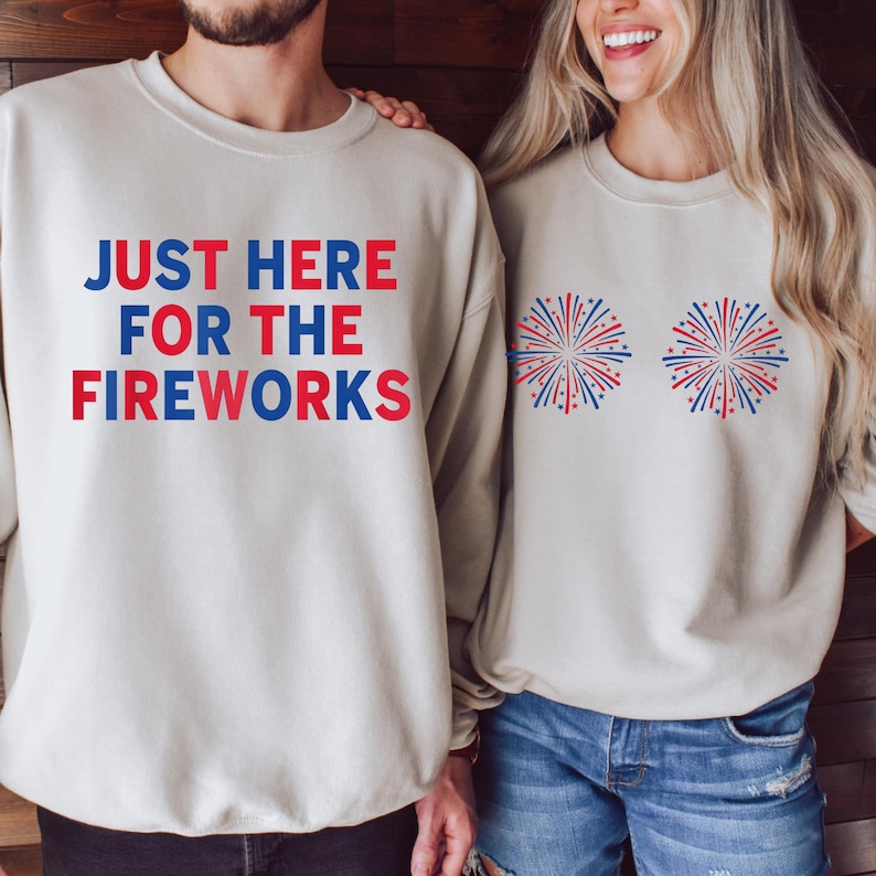 May include: Two beige crewneck sweatshirts. The left sweatshirt features the text "JUST HERE FOR THE FIREWORKS" in alternating red and blue block letters. The right sweatshirt features two graphic illustrations of red and blue fireworks.