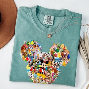 May include: A seafoam green t-shirt featuring a large Mickey Mouse head design composed of various Disney characters. The shirt has a crew neck and short sleeves. A silver necklace with a small pendant rests on the shirt.
