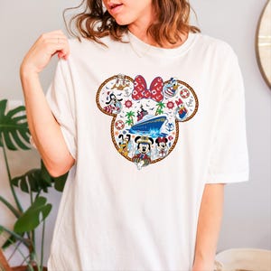 May include: White t-shirt with a Minnie Mouse-shaped design incorporating nautical themes. The design includes a cruise ship, Disney characters, and a red bow. The design is framed by a rope-style border.