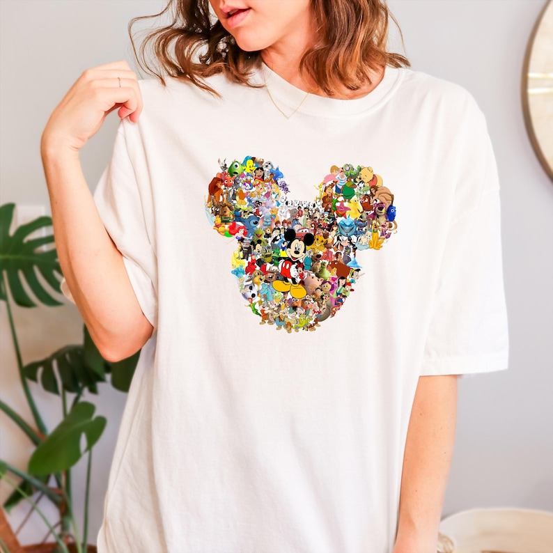 May include: White t-shirt featuring a colorful collage of Disney characters arranged in the shape of Mickey Mouse's head. The design includes characters like Mickey, Minnie, and Donald Duck.