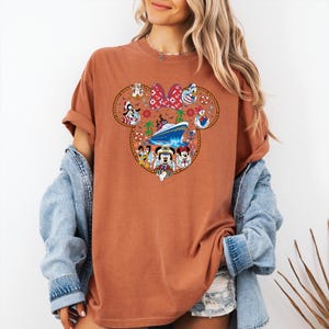May include: A rust-coloured t-shirt with a Mickey Mouse head design, featuring a cruise ship and Disney characters. The design includes Mickey, Minnie, Donald, and Goofy, surrounded by nautical elements. The shirt has short sleeves and a relaxed fit.