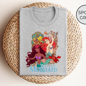 May include: Sport grey t-shirt featuring a colorful graphic of characters from "The Little Mermaid." The design includes Ariel, Flounder, Sebastian, and other characters. The words "THE LITTLE MERMAID" are printed in blue below the graphic.
