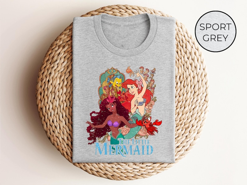 May include: Sport grey t-shirt featuring a colorful graphic of characters from "The Little Mermaid." The design includes Ariel, Flounder, Sebastian, and other characters. The words "THE LITTLE MERMAID" are printed in blue below the graphic.
