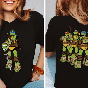 May include: Black t-shirt featuring a full-color graphic of Leonardo from Teenage Mutant Ninja Turtles on the front. The back of the shirt displays the four turtles. The design includes green, yellow, brown, and blue colors.