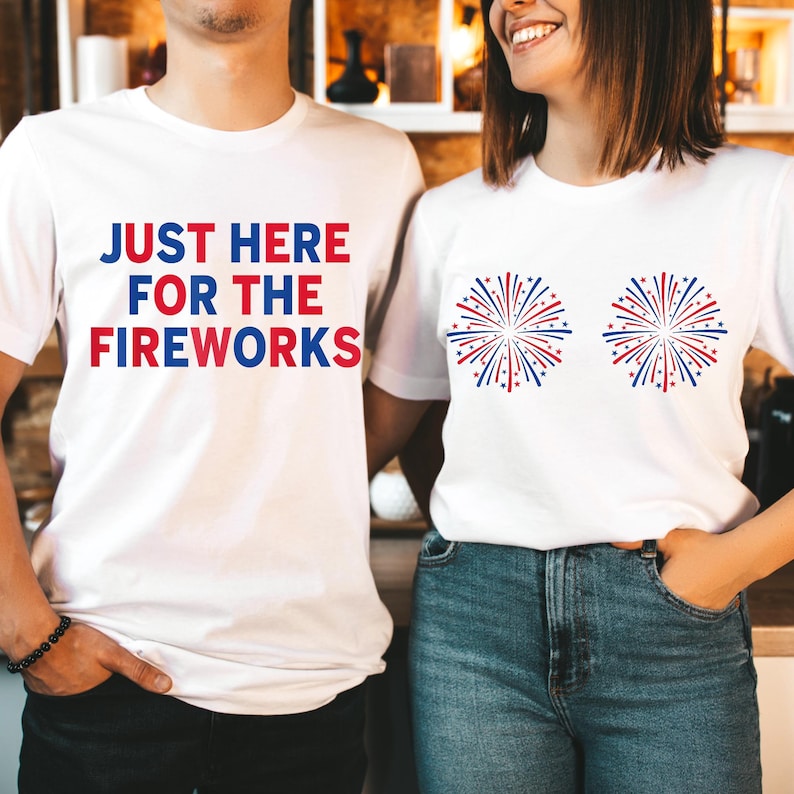 May include: Two white t-shirts. The left shirt features the blue and red text "JUST HERE FOR THE FIREWORKS". The right shirt features two graphic illustrations of red, white, and blue fireworks in a starburst pattern on the chest area.