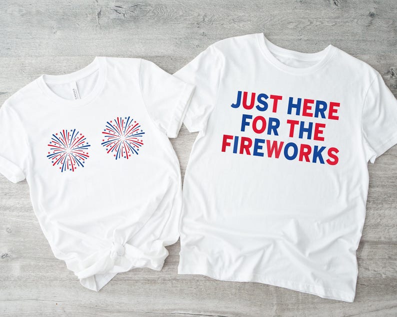 May include: Two white t-shirts. One shirt has the text "JUST HERE FOR THE FIREWORKS" in red and blue. The other shirt has two red, white, and blue firework designs.