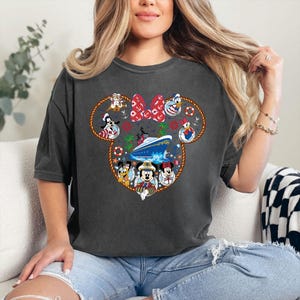 May include: Dark grey t-shirt with a Minnie Mouse-shaped design, featuring Disney characters and a cruise ship. The design includes a red bow, anchors, and palm trees. The shirt is made of a soft material.