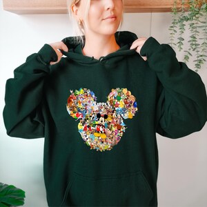 May include: Dark green hoodie featuring a Mickey Mouse head design composed of various cartoon characters. The hoodie has a front pocket and a drawstring hood. The design includes characters from Disney movies and shows.