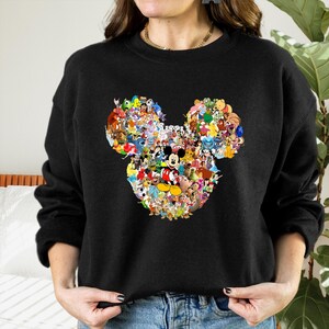 May include: Black sweatshirt featuring a large Mickey Mouse head design. The head is composed of a collage of various Disney characters in vibrant colors. Mickey Mouse is prominently displayed in the center, surrounded by other beloved characters.