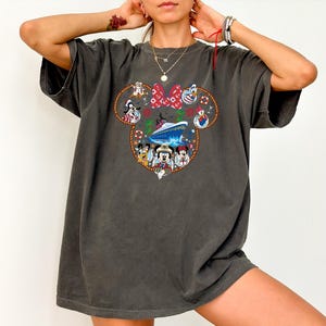May include: Dark grey t-shirt with a Mickey Mouse head design, showcasing Disney characters and a cruise ship. The design includes Minnie Mouse's bow, Donald Duck, and other characters. The shirt has short sleeves and a relaxed fit.