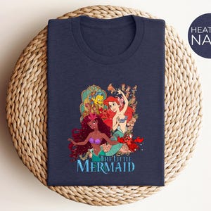 May include: Heather navy t-shirt featuring a colorful graphic of characters from "The Little Mermaid." The design includes Ariel, Flounder, Sebastian, and other mermaids. The words "The Little Mermaid" are printed in a blue font below the characters.