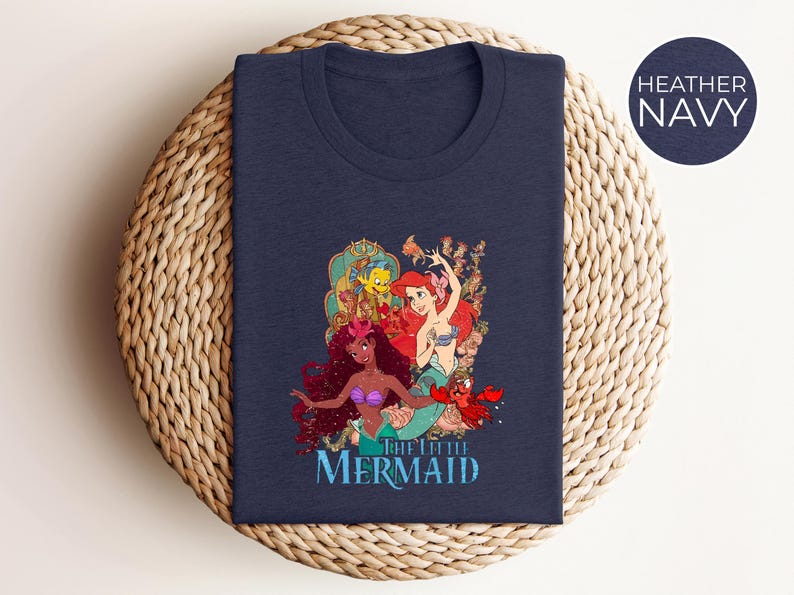May include: Heather navy t-shirt featuring a colorful graphic of characters from "The Little Mermaid." The design includes Ariel, Flounder, Sebastian, and other mermaids. The words "The Little Mermaid" are printed in a blue font below the characters.