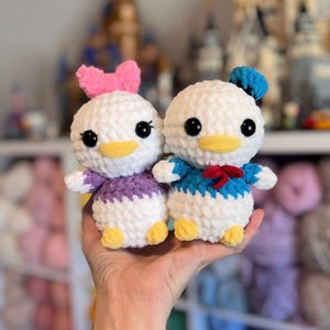 May include: Two handmade crochet dolls, one resembling Daisy Duck with a pink bow and purple outfit, and the other Donald Duck with a blue sailor shirt and hat. Both dolls have yellow beaks and feet, and black eyes.