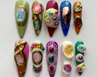 May include: Ten long, almond-shaped false nails with different designs. Each nail is decorated with a variety of colorful gems, glitter, and gold accents. The nails are arranged in two rows of five.