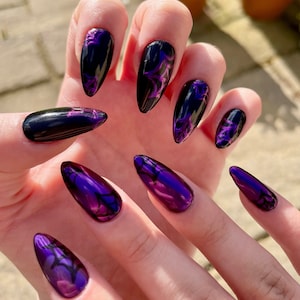 May include: Close-up of a hand with several long, almond-shaped nails painted with a dark purple and black nail polish. The nails have a swirling, marbled design.