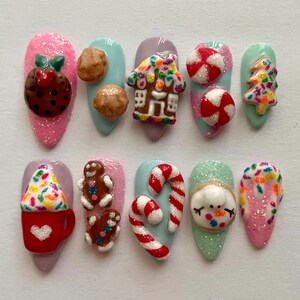 May include: A collection of ten artificial nails, each decorated with a festive Christmas design. The designs feature a gingerbread house, candy canes, a snowman, and a Christmas tree. The nails are painted in shades of pink, blue, and green.