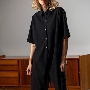 Könnte beinhalten: Ein schwarzer Jumpsuit mit kurzen Ärmeln, einem Kragen, einer durchgehenden Knopfleiste mit hellen Knöpfen und weit geschnittenen, verkürzten Hosenbeinen. Das Kleidungsstück besteht aus strukturiertem Stoff und hat eine lockere Passform.