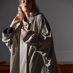 Puede incluir: Un impermeable beige claro de gran tamaño con capucha. El abrigo tiene una parte delantera abotonada, bolsillos y mangas largas. La persona de la imagen lleva el abrigo y tiene la mano cerca de la cara. El abrigo parece estar hecho de un material ligero.