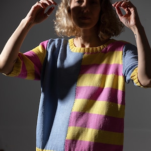 Op de afbeelding: Een colorblock trui met een blauw, geel en roze ontwerp. De trui heeft een ronde hals en korte mouwen. De linkerkant is effen blauw, terwijl de rechterkant horizontale gele en roze strepen heeft.