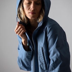Könnte beinhalten: Blaue Kapuzenjacke mit durchgehendem Reißverschluss und zwei Vordertaschen. Die Jacke ist aus leichtem Stoff gefertigt und hat eine Kapuze mit Kordelzug. Das Model trägt die Jacke.