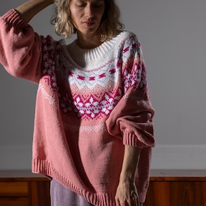 Op de afbeelding: Een roze trui met een wit en roze patroon over de borst en schouders. De trui heeft lange mouwen en een geribbelde halslijn en manchetten. Het model draagt een paarse corduroy broek.