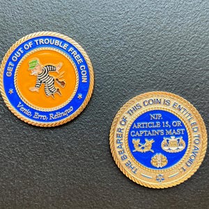Puede incluir: Dos monedas doradas con esmalte azul y naranja. Una moneda presenta un dibujo animado de un prisionero huyendo, con el texto "GET OUT OF TROUBLE FREE COIN". La otra moneda tiene el texto "THE BEARER OF THIS COIN IS ENTITLED TO AVOID..."