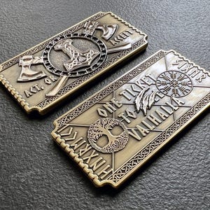 May include: Two gold-colored metal ticket replicas with intricate designs. One ticket reads "One Ticket To Valhalla" with a tree of life and runes. The other ticket reads "Act. Or Accept" with axes and a hammer.