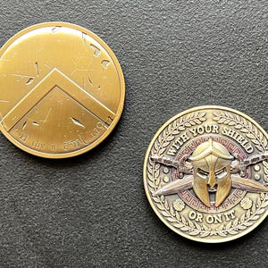 Könnte beinhalten: Zwei bronzefarbene spartanische Münzen. Eine Münze zeigt ein Schilddesign mit griechischem Text. Die andere Münze hat einen spartanischen Helm, gekreuzte Schwerter und den Text "WITH YOUR SHIELD OR ON IT".