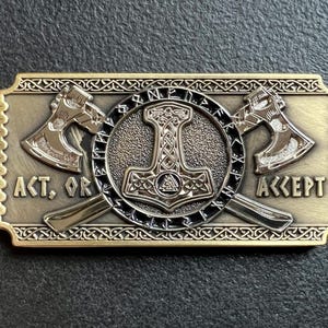 May include: A gold-toned metal belt buckle with a detailed design featuring two axes crossed behind a hammer with a knotwork design. The buckle has the words "ACT, OR ACCEPT" inscribed on it.