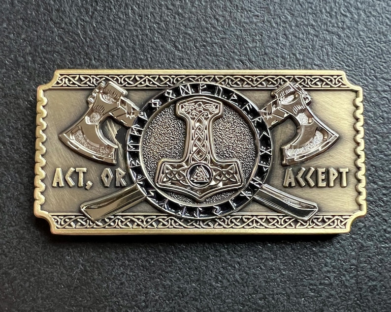 May include: A gold-toned metal belt buckle with a detailed design featuring two axes crossed behind a hammer with a knotwork design. The buckle has the words "ACT, OR ACCEPT" inscribed on it.