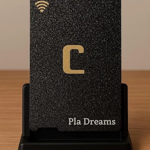 Puede incluir: Una tarjeta negra con superficie texturizada, con la letra "C" dorada y un símbolo Wi-Fi. El texto "Pla Dreams" está impreso en la parte inferior. La tarjeta se muestra en un soporte negro sobre una superficie de madera.