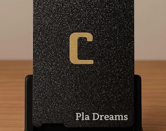Op de afbeelding: Een zwarte kaart met een getextureerd oppervlak, met de gouden letter "C" en een Wi-Fi-symbool. De tekst "Pla Dreams" staat onderaan gedrukt. De kaart staat in een zwarte standaard op een houten oppervlak.