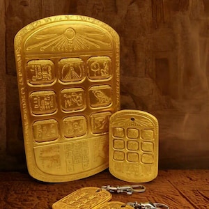 May include: A collection of gold-colored rectangular plaques and keychains featuring embossed Egyptian-style hieroglyphics and symbols. The items include a large plaque, a smaller plaque with a hole at the top, and two keychains with metal clips.