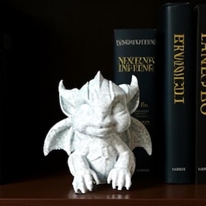 Puede incluir: Una pequeña estatua de gárgola gris claro con alas, cuernos y garras detalladas. La estatua está colocada sobre una estantería de madera, con libros oscuros en el fondo. La gárgola tiene una expresión serena.