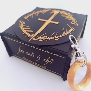 May include: Black box with gold accents, featuring a sword and inscription. A gold ring is attached via a silver clasp. The box has the text "The way is shut" in gold lettering. The ring is a light orange color.
