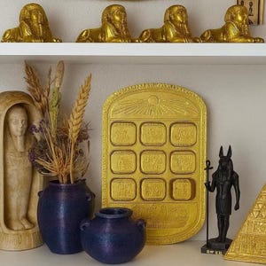 May include: A shelf displays several golden sphinx statues, a golden tray with rectangular compartments, and a black statue of Anubis. Two blue vases hold dried flowers, and a carved figure is also present.