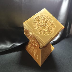 Peut inclure: Un cube imprimé en 3D de couleur dorée avec des motifs complexes sur chaque face, reposant sur une base pyramidale. Le cube présente un motif circulaire avec une figure centrale et des motifs géométriques.