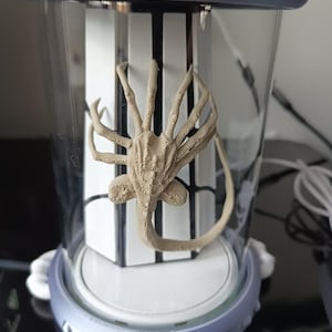 May include: A tan-colored alien facehugger figurine inside a clear cylindrical container with a light blue top and base. The container has a white and black striped background. The figurine has a long tail and multiple legs.