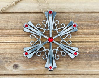 Puede incluir: Un adorno con forma de copo de nieve hecho de clavos de metal plateado dispuestos en un patrón radial. El centro y cada una de las seis puntas están decorados con una pequeña piedra preciosa roja brillante. Incluye un lazo de cuerda fina.