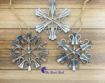 Puede incluir: Tres adornos en forma de copo de nieve hechos de clavos de herradura de metal plateado, cada uno con un punto central con brazos radiantes y extremos curvados. Cada adorno incluye un lazo de cuerda de color canela para colgarlo.