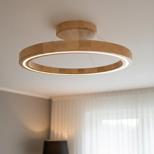 Könnte beinhalten: Eine moderne, kreisförmige Deckenleuchte aus Holz. Die Leuchte hat eine warme, natürliche Holzoberfläche und eine helle, integrierte LED-Lichtquelle, die das Innere des Rings beleuchtet. Die Leuchte wird mit dünnen Drähten von der Decke abgehängt.