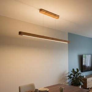 Puede incluir: Una lámpara colgante de madera de diseño moderno. Está suspendida del techo por finos cables e ilumina una mesa de comedor. La lámpara es de madera clara y de forma rectangular, con una luz lineal.