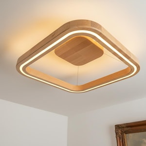 Può includere: Lampada da soffitto quadrata in legno con finitura in legno naturale e calda. La lampada ha una cornice quadrata con una striscia illuminata incassata e un pannello centrale quadrato. Montata su un soffitto bianco.