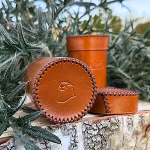 May include: A cylindrical tan leather case with a matching lid, featuring dark brown cross-stitch detailing along the edges and a small embossed beaver head logo on the circular base. The leather surface is smooth with a warm, light brown tone.