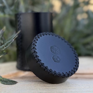 Puede incluir: Vaso de dados de cuero negro con tapa a juego. La tapa presenta dos símbolos de dados y está cosida alrededor del borde. La copa es cilíndrica y también tiene detalles de costura. El fondo está borroso, con follaje verde y una superficie de madera.