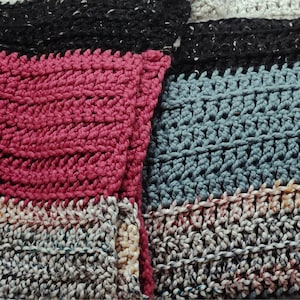 May include: A close-up of a multi-colored crocheted blanket with different patterns and textures. The blanket is made with a variety of yarn colors, including pink, blue, black, and white.