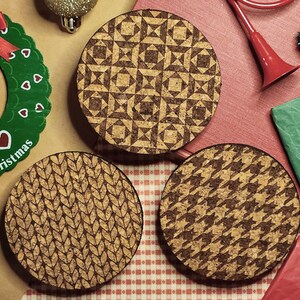 May include: Three round cork coasters with different geometric patterns. The coasters are brown with black designs. The patterns include a quilt block design, a herringbone pattern, and a knit pattern.