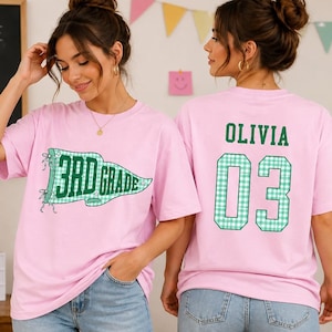 May include: A light pink short-sleeve t-shirt featuring a green and white checkered pennant graphic on the front with the text 3RD GRADE. The back of the shirt displays the name OLIVIA in green letters above the large green and white checkered number 03.