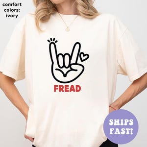 Puede incluir: Camiseta de color marfil con un gesto de la mano negro, un corazón y la palabra "FREAD" en rojo. La camiseta tiene un corte holgado y está hecha de un material suave. El texto "comfort colors: ivory" está en la esquina superior izquierda.